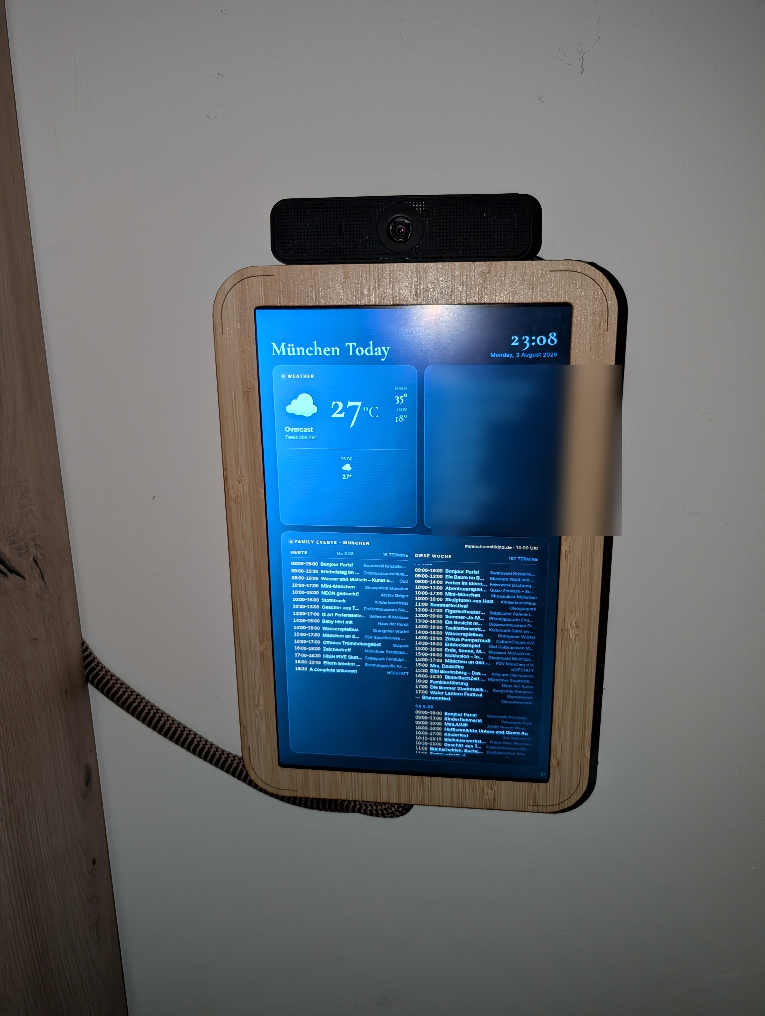 The framed X201 screen mounted on a wall, showing a family dashboard with weather and local events (personal calendar blurred)